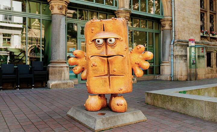 A statue of a loaf of bread with hands, feet and a grim face stands in front of a building with many glass windows.