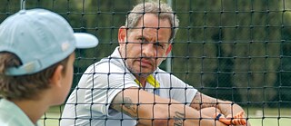 The picture shows actor Moritz Bleibtreu in a scene of the film Everything's Fifty Fifty by Alireza Golafshan. He sits behind a tennis net and looks at a child. The child is close, blurry and only seen from behind. 