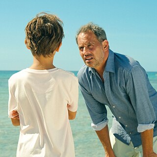The picture shows actor Moritz Bleibtreu in the film Everything's fifty fifty by Alireza Golafshan. He is standing on the beach, ocean behind him. He leans forward, hands on this knees and looks at a boy. The boy is only seen from behind. 