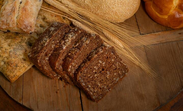 Different types of bread on a wooden base. In the centre, a sliced dark loaf with lots of grains.