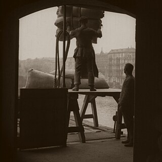 Ausschnitt aus dem Film Buddenbrooks, in dunklen Sepia-Tönen. Eine Person nimmt auf einer Hebebühne schwere Säcke entgegen, eine andere schaut dabei zu.