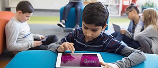 Ein Junge benützt in einer Bibliothek ein Tablet. Im Hintergrund weitere lesende Kinder mit Tablets.