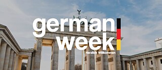 The picture shows a photo of the Brandenburger Tor in Berlin. In the background the sky with a few clouds. In the foreground is a writing that reads "German Week". Next to the writing on the right hand side is a small black-red-gold streak.