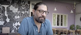 Interview mit Ghassan Sahhab vor einer kunstvoll gestalteten Wand mit grafischen Elementen. Im Hintergrund sind Tische, Stühle und grüne Pflanzen zu sehen.