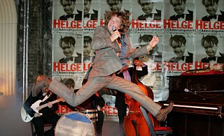 A white man in a suit with a microphone in his hand jumps, behind him a band and posters with the words ‘Helge, Admiralspalast’.