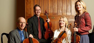 A string quartet poses with their instruments in a room with wooden walls.