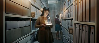 A young woman is standing between archiv shelfs and looks at a small apparition of a couple dancing
