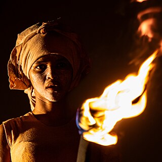 GPS Project - Ons Gister Daans: a person with a cloth wrapped around the head, holding a torch with a bright flame. The background is dark, highlighting the illumination from the torch.