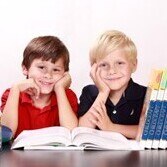 Zwei Grundschulkinder sitzen lächelnd am Schreibtisch und haben ihre Ellenbogen aufgestützt. Links neben ihnen steht ein Globus und rechts stehen ein paar Bücher.