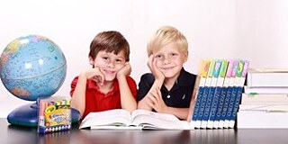 Zwei Grundschulkinder sitzen lächelnd am Schreibtisch und haben ihre Ellenbogen aufgestützt. Links neben ihnen steht ein Globus und rechts stehen ein paar Bücher.