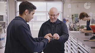 Zwei Personen stehen in einem Werkraum und tauschen eine Schutzbrille aus.