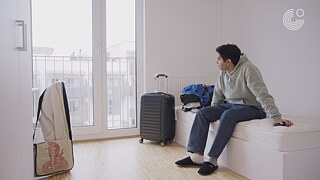 Eine Person sitzt auf einem Bett in einem Zimmer, neben einem Koffer und Kleidung, mit Blick auf das Fenster