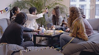 Mehrere Personen sitzen in einem Wohnzimmer um einen gedeckten Tisch und stoßen gemeinsam an, umgeben von Geburtstagsdekoration.