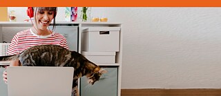 Una ragazza con un portatile e una cuffia rossa sorride mentre un gatto le passa sulla tastiera