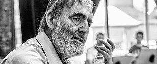 A man facing the viewer in the foreground, musicians in the background.