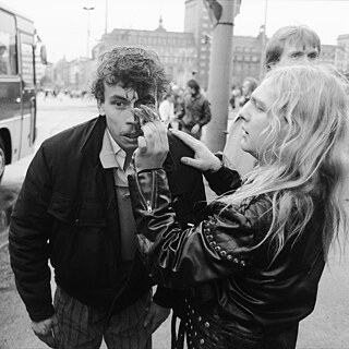 Das Foto zeigt einen Mann in Lederjacke und mit langen Haaren, der einem anderen Mann mithilfe eines Tuches  Blut aus dem Gesicht entfernt. Im Hintergrund sieht man eine große Straße und einen Bus.