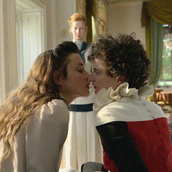 To kvinder kysser hinanden, en tredje står i baggrunden og ser på.