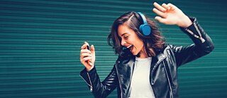 A woman with brown hair wearing blue headphones moves to the music.  She holds an orange cell phone in her left hand.  Her mouth is open as if she is singing along, and her eyes are closed.