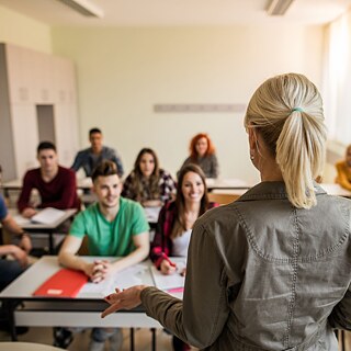 Rückansicht auf eine Lehrerin, die zu Schülern der Sekundarstufe im Klassenraum spricht.
