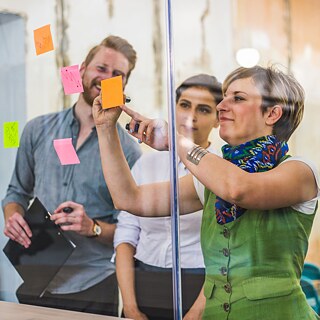 Drei Teamkollegen arbeiten kreativ mit Post-its an einer Glaspinnwand. Sie sind sehr konzentriert und haben Spaß.