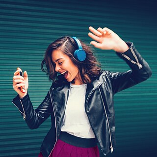 A woman with brown hair wearing blue headphones moves to the music.  She holds an orange cell phone in her left hand.  Her mouth is open as if she is singing along, and her eyes are closed.
