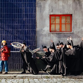 Eine Frau in roter Jacke und gelber Mütze geht an einer Gruppe von Männern in schwarzen Gewändern vorbei, die theatralisch gestikulieren und Fahrräder tragen.