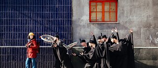 Eine Frau in roter Jacke und gelber Mütze geht an einer Gruppe von Männern in schwarzen Gewändern vorbei, die theatralisch gestikulieren und Fahrräder tragen.