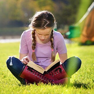 A girl sits cross-legged on a meadow and looks into a book lying on her legs. The weather is sunny.
