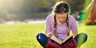 A girl sits cross-legged on a meadow and looks into a book lying on her legs. The weather is sunny.