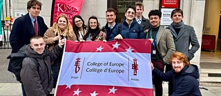 Eine Gruppe Studenten steht vor einem Gebäude und hält eine Fahne des College of Europe vor sich.