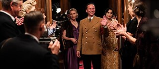 Ein Mann in brauner Uniformjacke schreitet über den roten Teppich und winkt dem applaudierenden Publikum zu, während Frauen in eleganten Kleidern und Kameras im Hintergrund die Szene begleiten