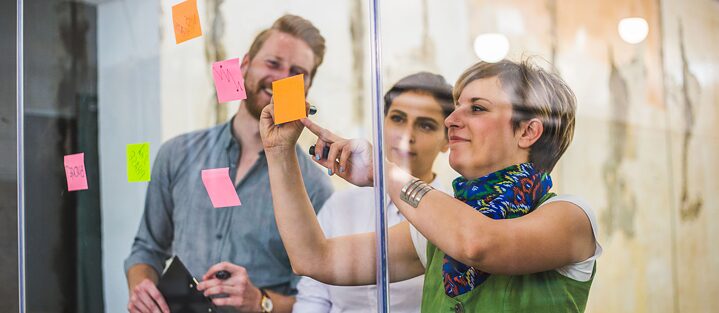 Drei Teamkollegen arbeiten kreativ mit Post-its an einer Glaspinnwand. Sie sind sehr konzentriert und haben Spaß.