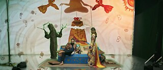 Un hombre está sentado descalzo con una shisha sobre un podio, a su lado una mujer con un vestido colorido, acompañados por una figura con forma de árbol, mientras sobre ellos flotan pájaros y una silueta en meditación