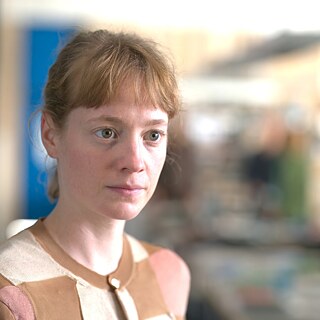 Eine Person mit rötlich-blonden Haaren und hellem Pullover blickt ernst in die Kamera, im Hintergrund ist ein unscharfer Innenraum erkennbar