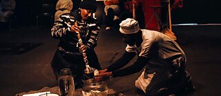 Tebello: Einer Performerin befüllt in einer rituellen Zeremonie das Gefäß eines Teilnehmers.