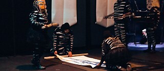 Tebello: Zwei Performende befestigen eine Rorschach-Malerei an einem Aufhängung, zwei Performende mit Masken stehen daneben und beobachten die Szene.