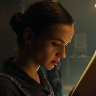 Eine Frau mit hochgesteckten Haaren liest konzentriert in einem Buch, während im Hintergrund eine Schale mit Obst und ein unscharfes Bett zu sehen sind