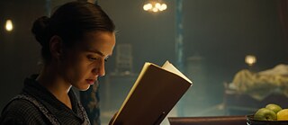 Eine Frau mit hochgesteckten Haaren liest konzentriert in einem Buch, während im Hintergrund eine Schale mit Obst und ein unscharfes Bett zu sehen sind