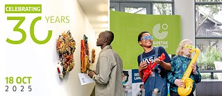 Das Bild ist in zwei Abschnitte unterteilt. Links befindet sich ein grünes und weißes Banner mit dem Text "CELEBRATING 30 YEARS" und "18 OCT 2025." Rechts interagieren drei Personen mit bunten Kunstwerken an einer Wand und spielen mit aufblasbaren Musikinstrumenten vor einem grünen Goethe-Institut-Banner.
