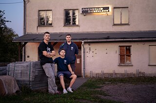 Three young people in sportswear are standing and sitting in front of an old, plastered building with a crumbling façade. Next to them are stacked paving stones.