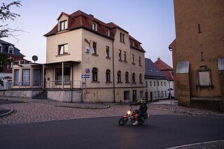 A motorcyclist wearing a yellow helmet rides through a quiet cobblestone street corner at dusk. Older buildings stand in the background, including a yellow house and a brown building with a commemorative plaque.