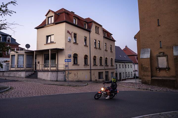 A motorcyclist wearing a yellow helmet rides through a quiet cobblestone street corner at dusk. Older buildings stand in the background, including a yellow house and a brown building with a commemorative plaque.