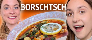 Ein Foto von zwei Frauen, die in die Kamera lachen. Zwischen ihnen ist eine Großaufnahme eines Suppentellers, der mit Eintopf gefüllt ist.