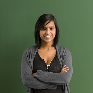 Eine junge Lehrerin steht mit verschränkten Armen vor einer grünen Tafel.