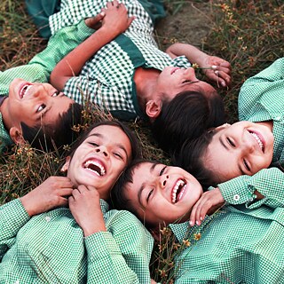 Eine Gruppe Schulkinder, in grünen Schuluniformen, liegt Kopf an Kopf im Gras und lacht. Close-Up, von oben auf die Gesichter der Kinder.