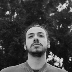 Auf dem Schwarz-Weiß Foto ist der Künstler Bruno Aguiar zu sehen. Das Foto zeigt sein Gesicht und er schaut nicht in die Kamera. Im Hintergrund befindet sich ein Baum, der aufgrund der Bearbeitung des Fotos eher unscharf ist. 