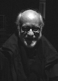 A fotografia a preto e branco mostra o artista Carlos «Zingaro». Ele usa óculos com armação preta e um casaco de inverno preto e sorri para a câmara.