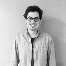 Das Schwarz-weiß Foto zeigt den Künstler Francisco Lima da Silva, der in die Kamera blickt und eine Brille trägt und vor einem weißen Hintergrund steht.