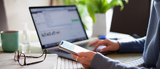 Una empresaria está sentada en casa frente al portátil y revisa los mensajes de texto en su teléfono inteligente. Delante de ella hay una taza de café.