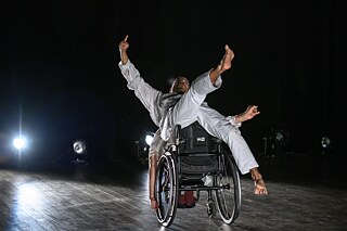 Un bailarín en una silla de ruedas realiza un movimiento de danza dinámico en un escenario. El bailarín lleva un atuendo gris y extiende los brazos y las piernas en diferentes direcciones. En el fondo se pueden ver varios focos que iluminan el espacio oscuro del escenario.